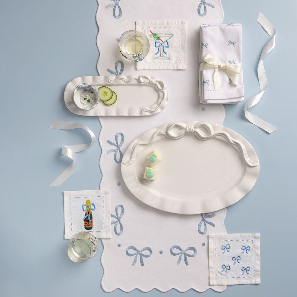 White cocktail napkins with blue embroidered bows, matching table runner, and bow-themed ceramic platters.