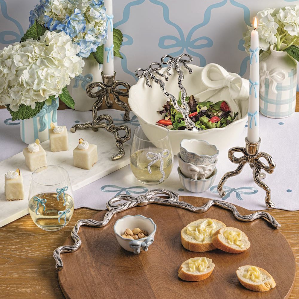 Wooden serving board with silver bow handles and appetizers on a table with bow-themed decor and blue accents.