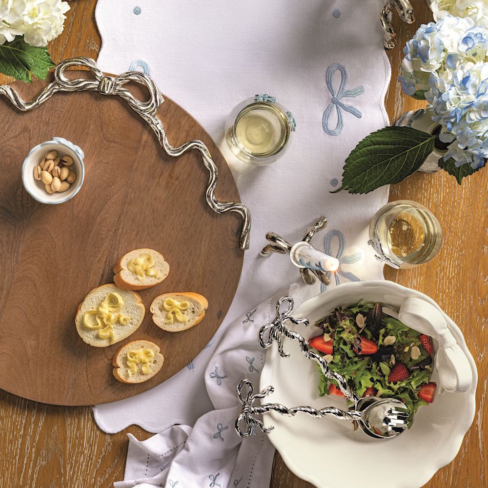Round wooden serving board with silver bow detailing, set on a table with appetizers, salad, and wine.
