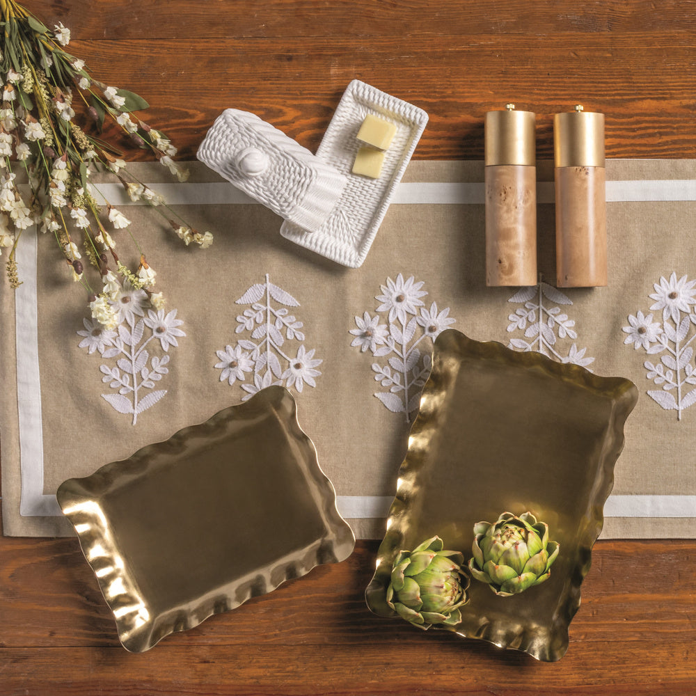 Two ruffled brass nesting trays on a floral table runner with a white butter dish, wood salt shakers, and artichokes.