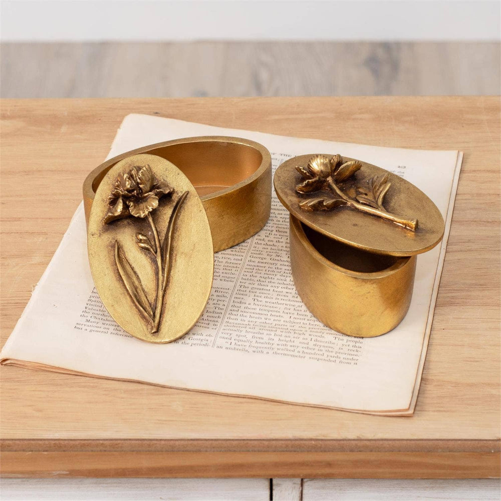 Two brass-tone oval floral trinket boxes with embossed iris and rose lids on vintage book pages.