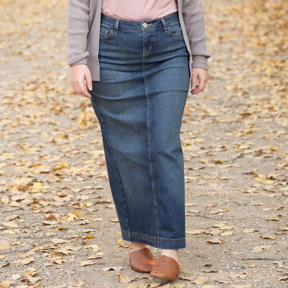 A woman wearing a long denim maxi skirt, cardigan, and brown leather mules on a path with autumn leaves.