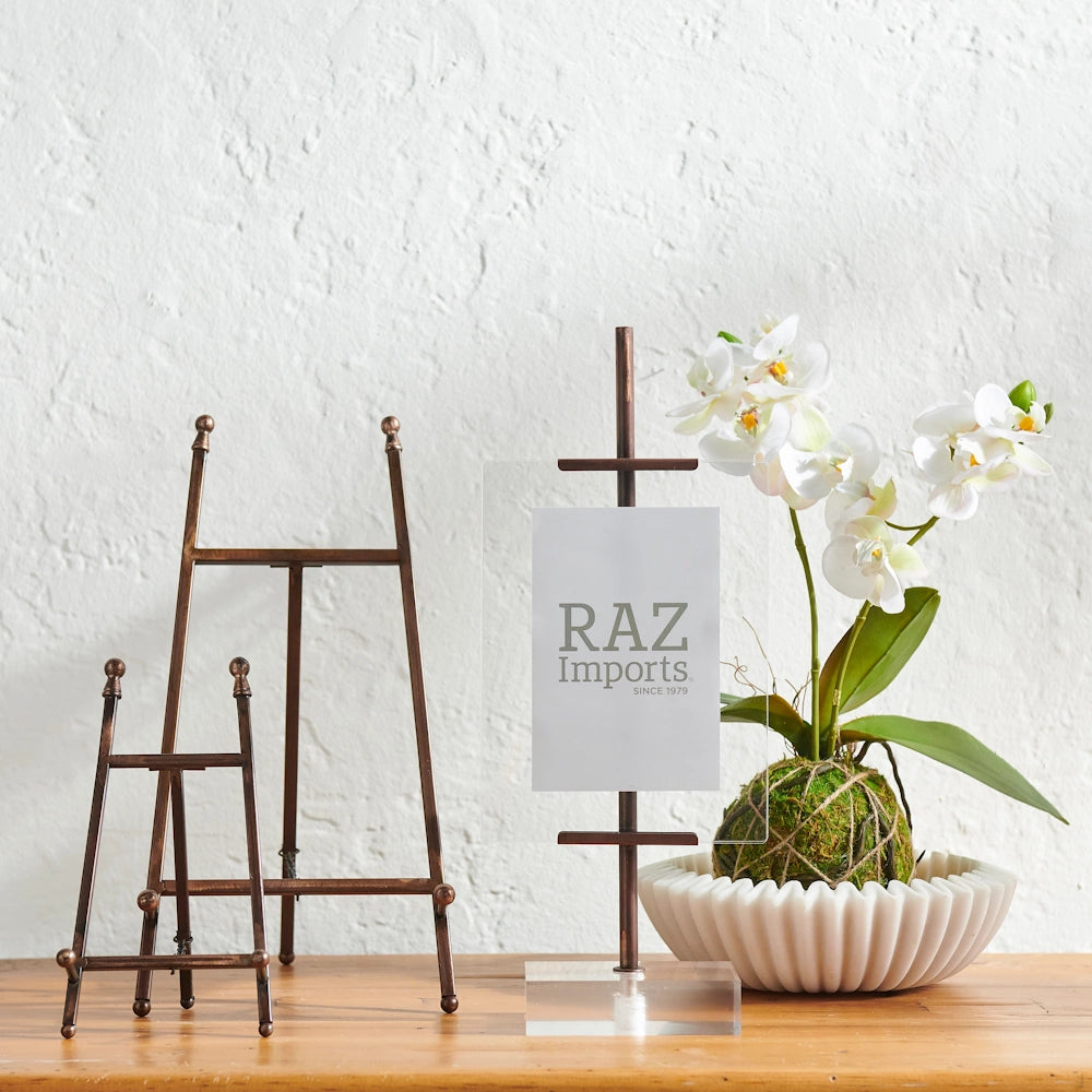 Two bronze-finished metal easels in different sizes next to a sign holder and a white orchid in a moss ball.