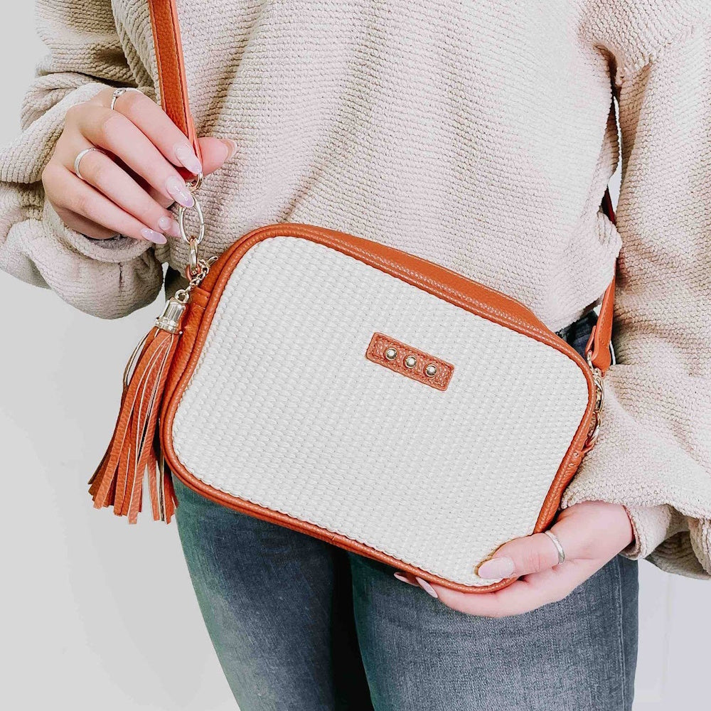 A person wearing a knit sweater and jeans holds a white woven crossbody bag with brown leather trim.