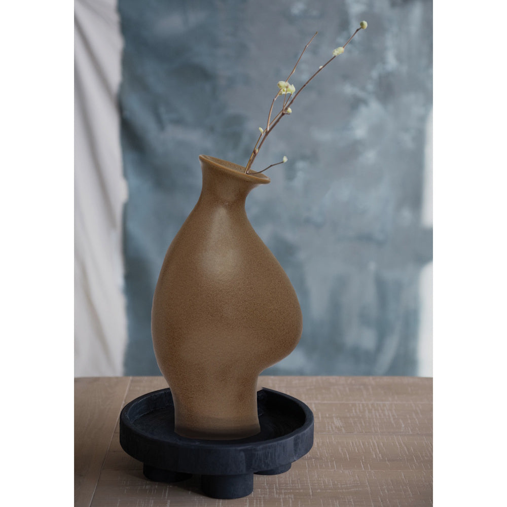 Organic-shaped brown ceramic vase holding a budded branch, set on a dark wooden tray against a textured wall.