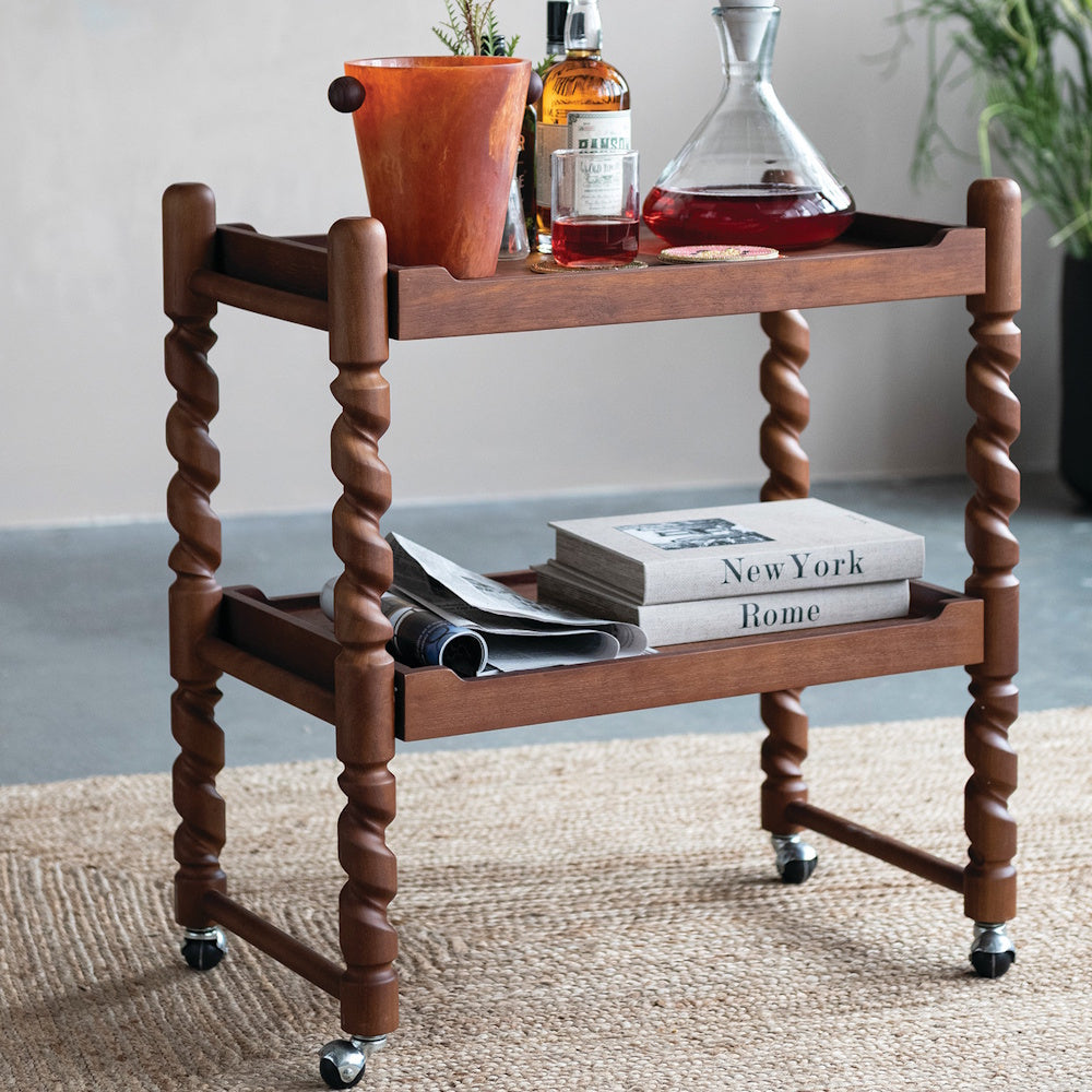 Two-tier brushed rubberwood bar cart with spiral legs, liquor bottles, glassware, and books on a woven rug.