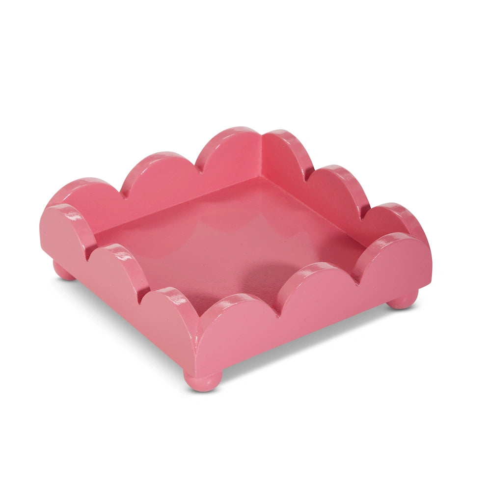 A square, bubblegum pink napkin holder with scalloped edges and small ball feet on a white background.