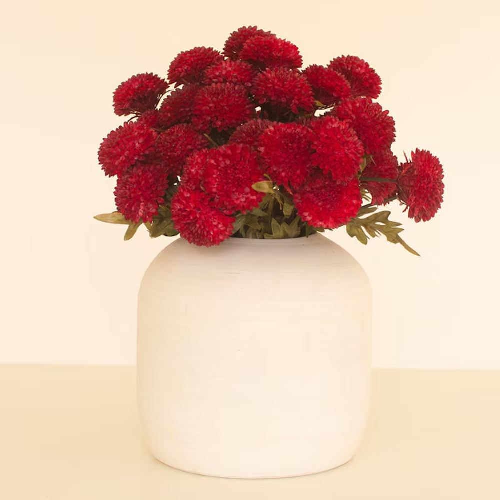 Artificial deep red ball dahlias arranged in a round white ceramic vase against a neutral background.