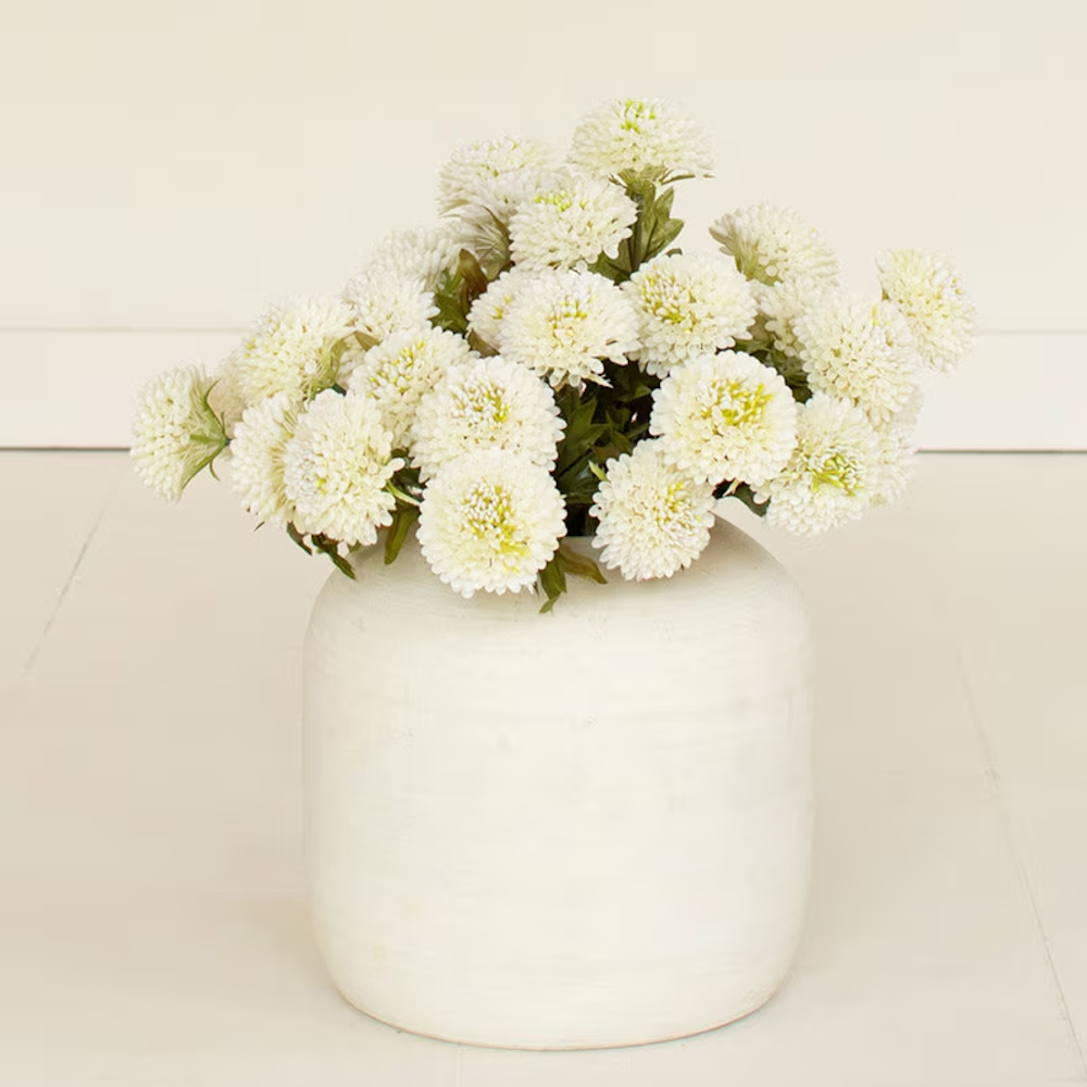 White artificial ball dahlias arranged in a round ceramic vase on a light backdrop.