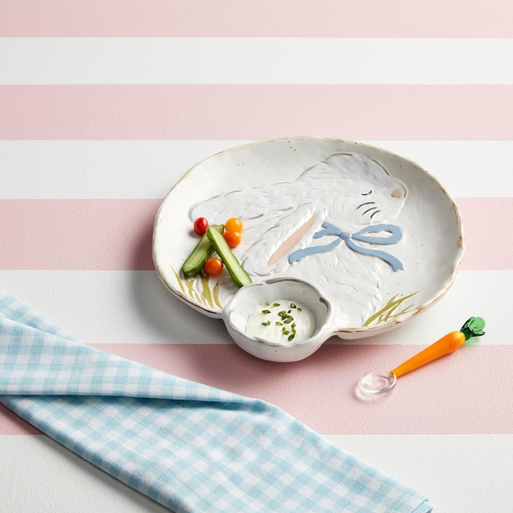 A bunny-shaped ceramic platter with vegetables, dip, and a carrot spoon on a pink striped surface.