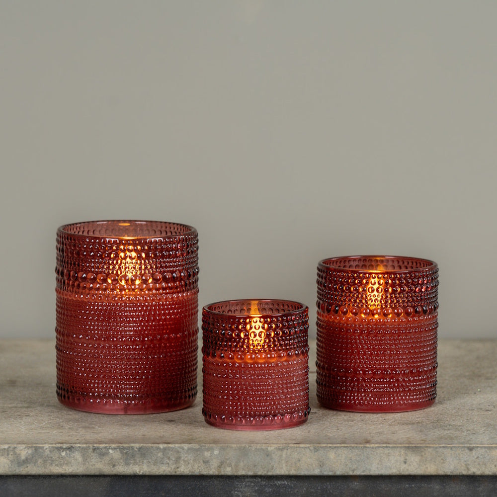 Three burgundy dotted glass votives with glowing 3D flameless candles in varying sizes on a stone ledge.