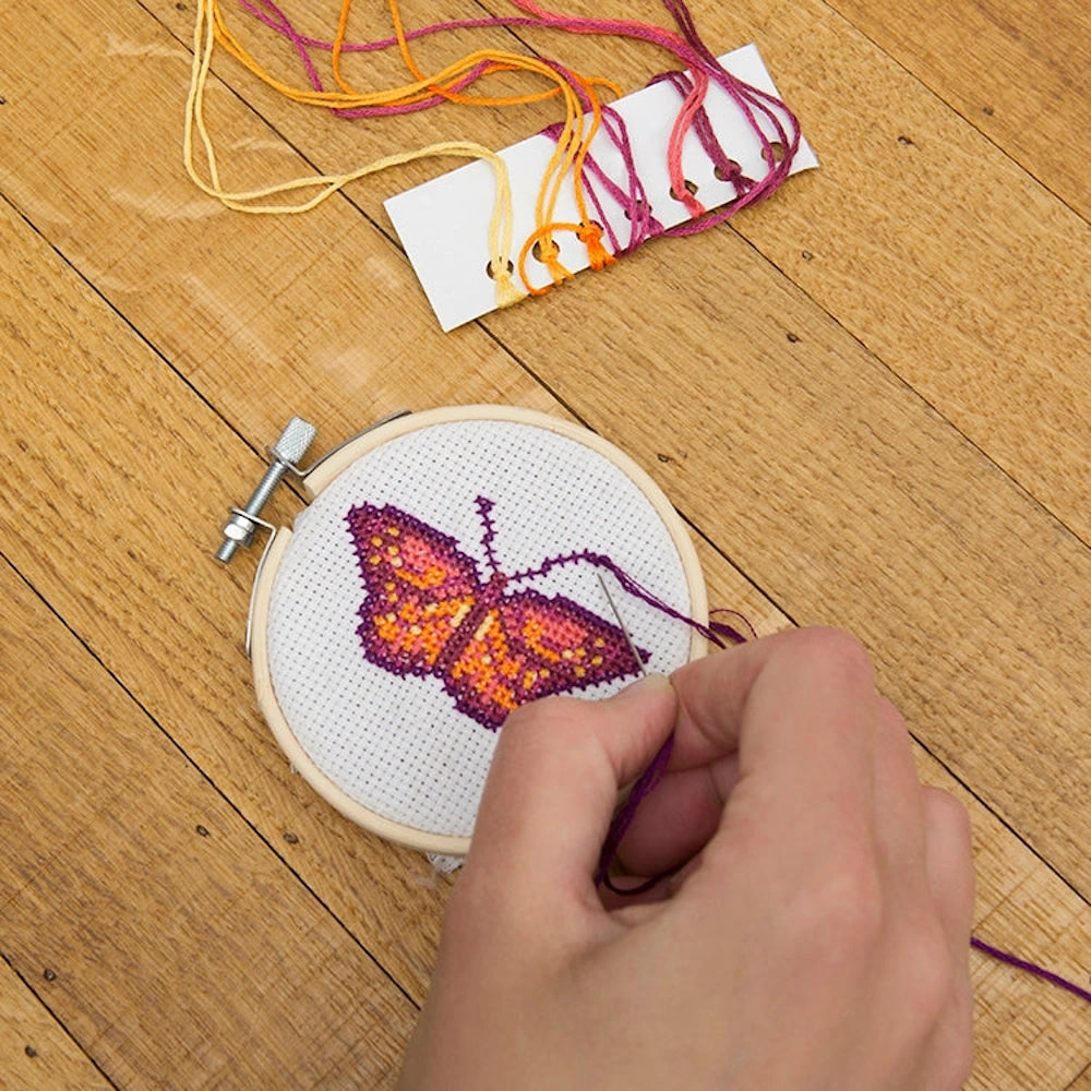 Hands stitching a purple and orange butterfly mini cross-stitch kit in a wooden hoop on a wood background.