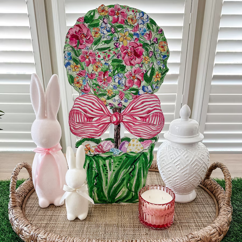 Easter floral topiary in a cabbage pot with pink bow, white bunnies, and a candle on a woven tray.