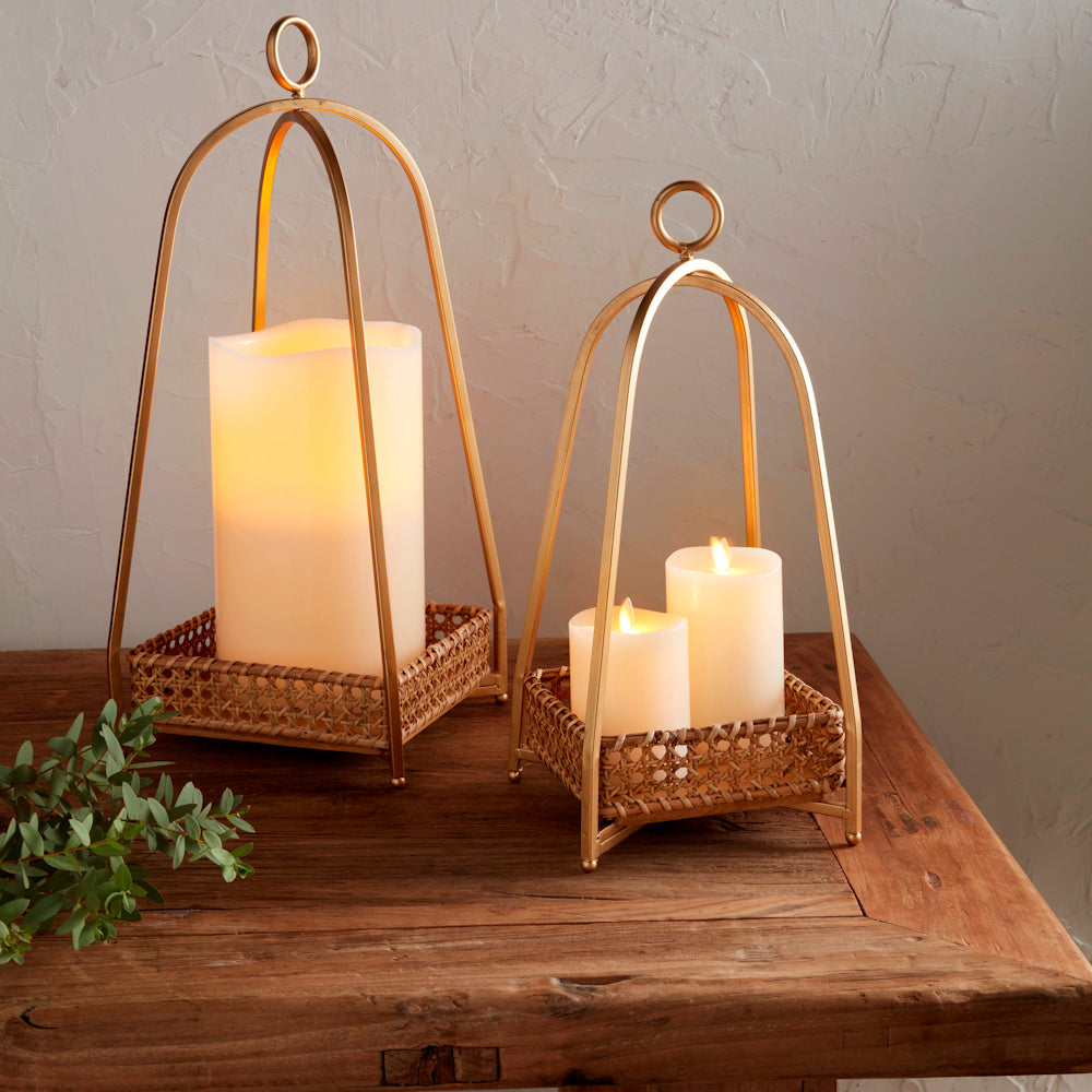 Two gold metal arch lanterns with woven cane trays and lit pillar candles on a rustic wooden table.