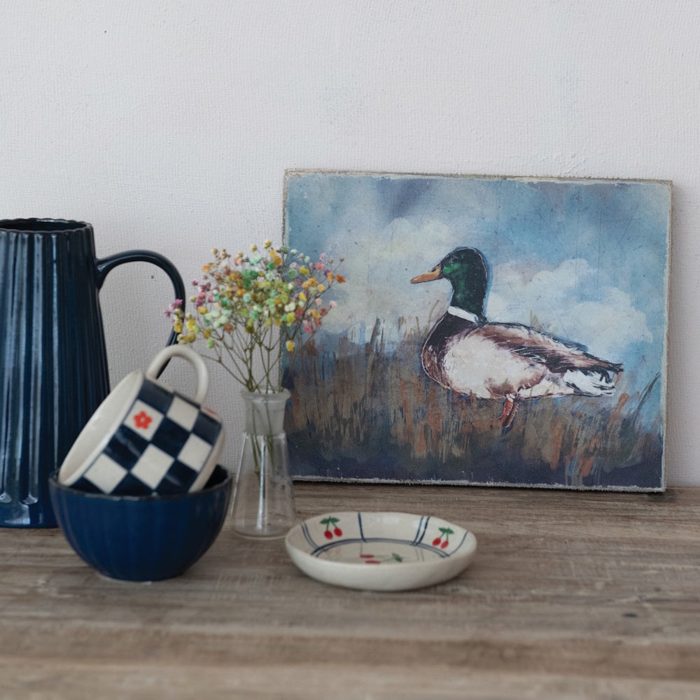 Mallard duck canvas wall art displayed on a rustic wooden table with ceramic dishware and small flowers.