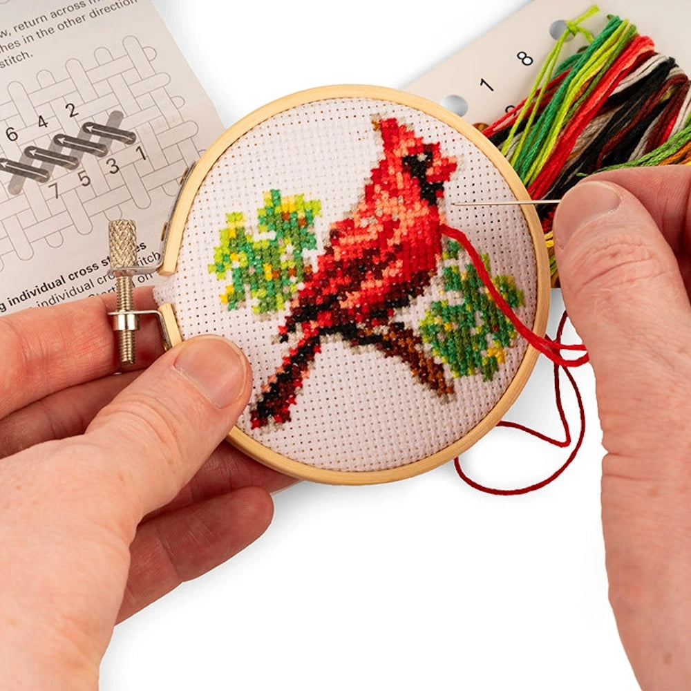 Mini cardinal cross-stitch embroidery kit featuring a red bird on a branch, held in a wooden hoop with thread and needle.