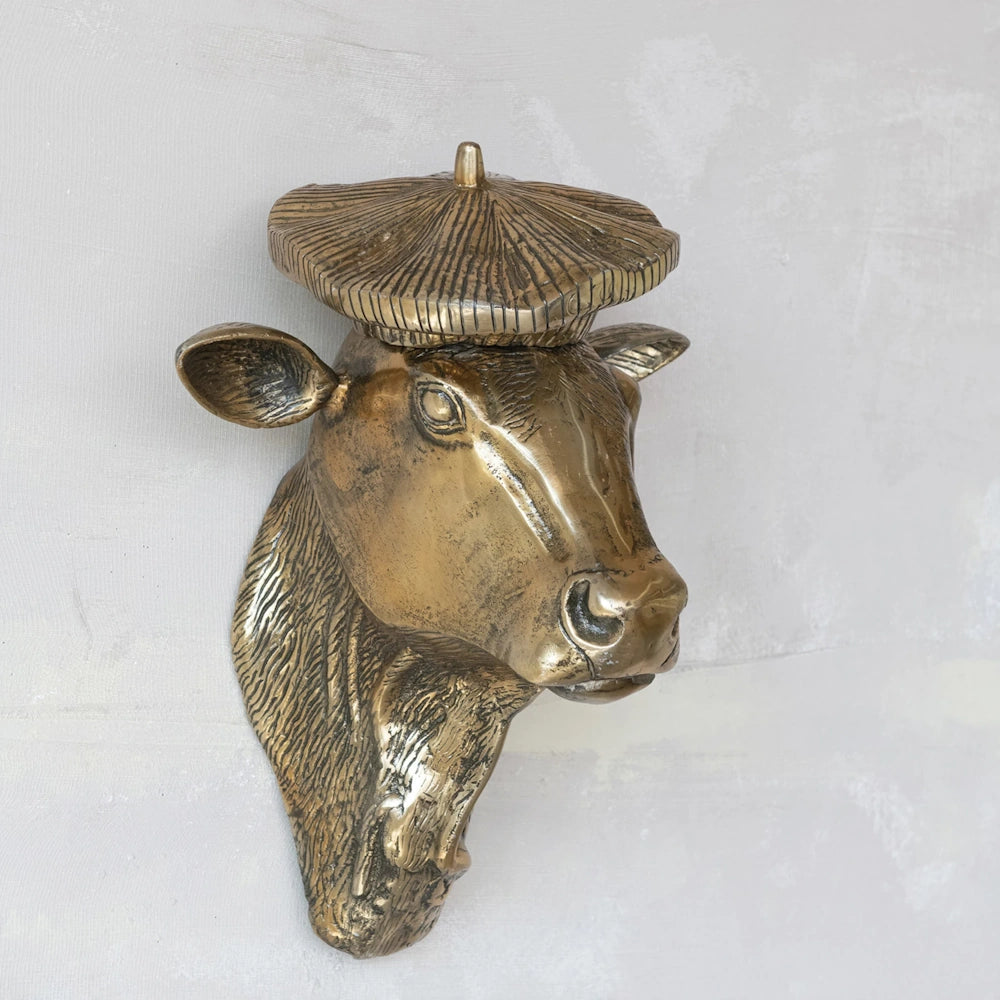 Bronze cow head wall sculpture wearing a beret, shown angled on a textured, light grey wall.