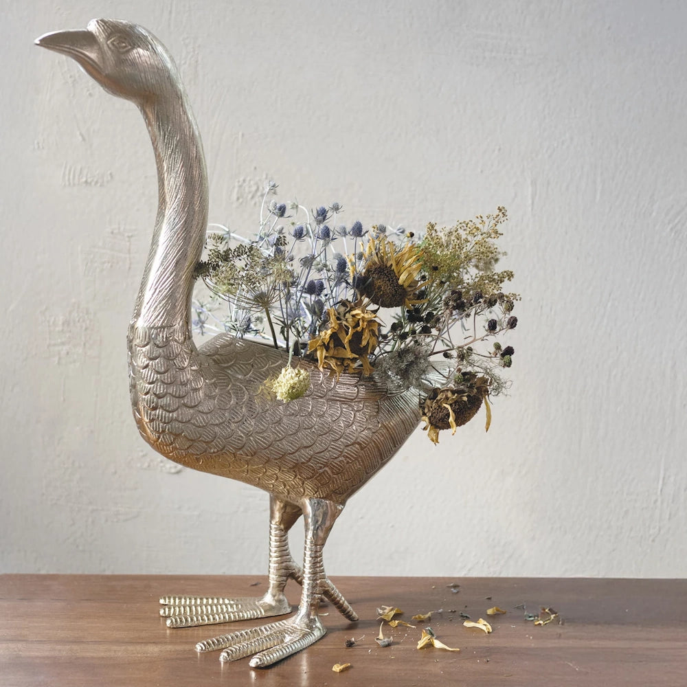 Silver cast aluminum goose-shaped planter filled with dried flowers on a wooden surface against a white wall.
