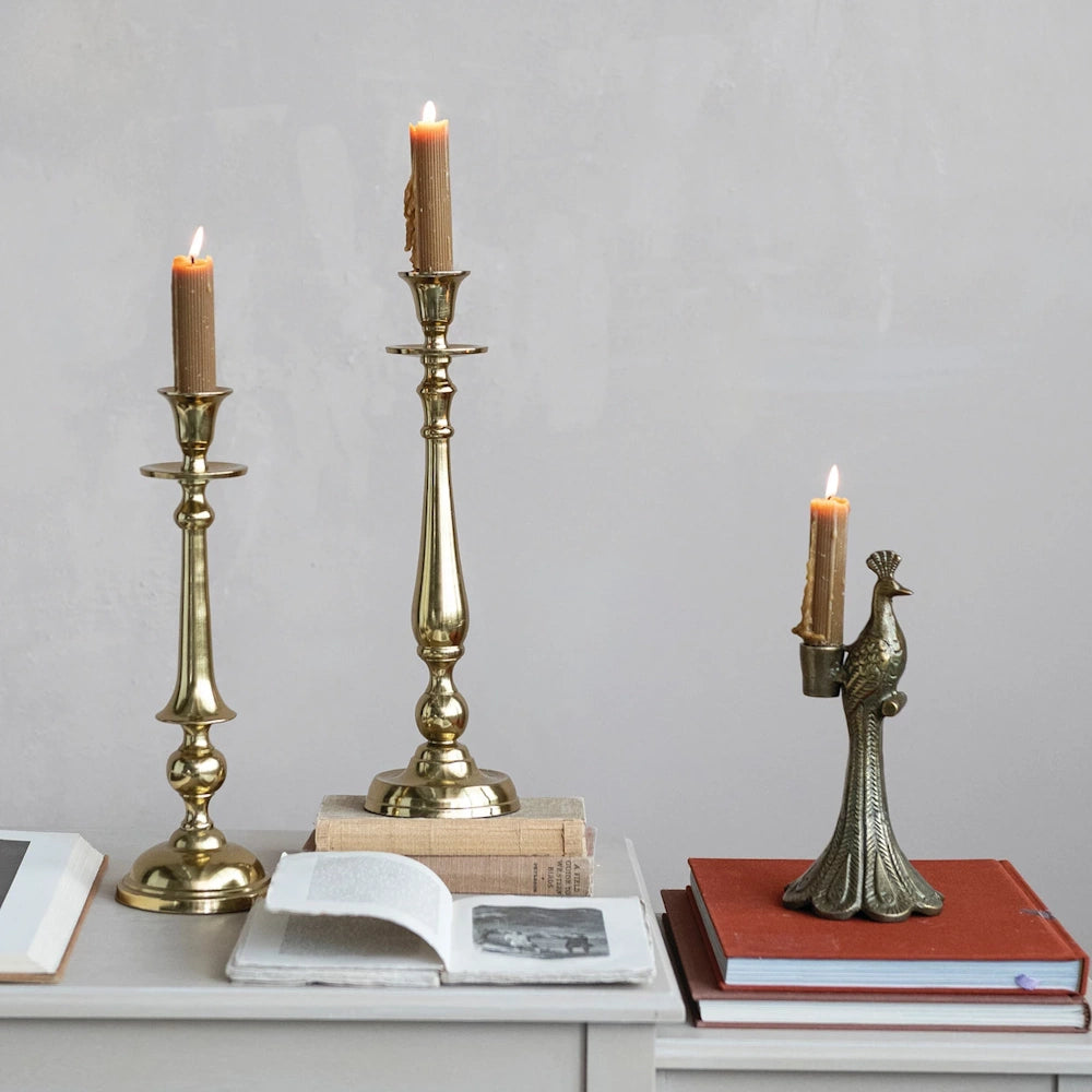 Antique peacock and brass taper candle holders with lit beeswax candles on a table with vintage books.