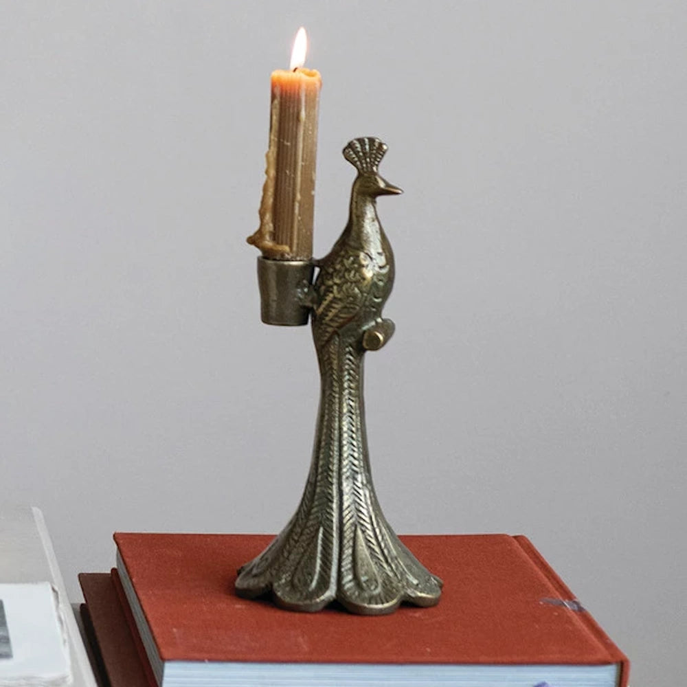 Antique-style cast aluminum peacock taper candle holder with a lit beeswax candle on a stack of books.