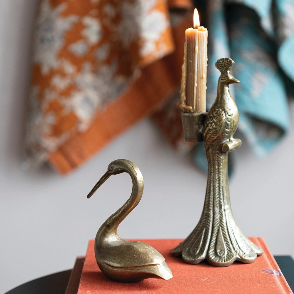 Vintage brass peacock candle holder and swan figurine on an orange book, featuring a lit taper candle and textile background.