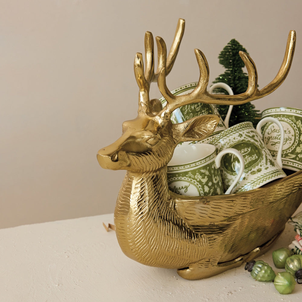 A gold cast aluminum reindeer-shaped bowl holding green and white holiday mugs.