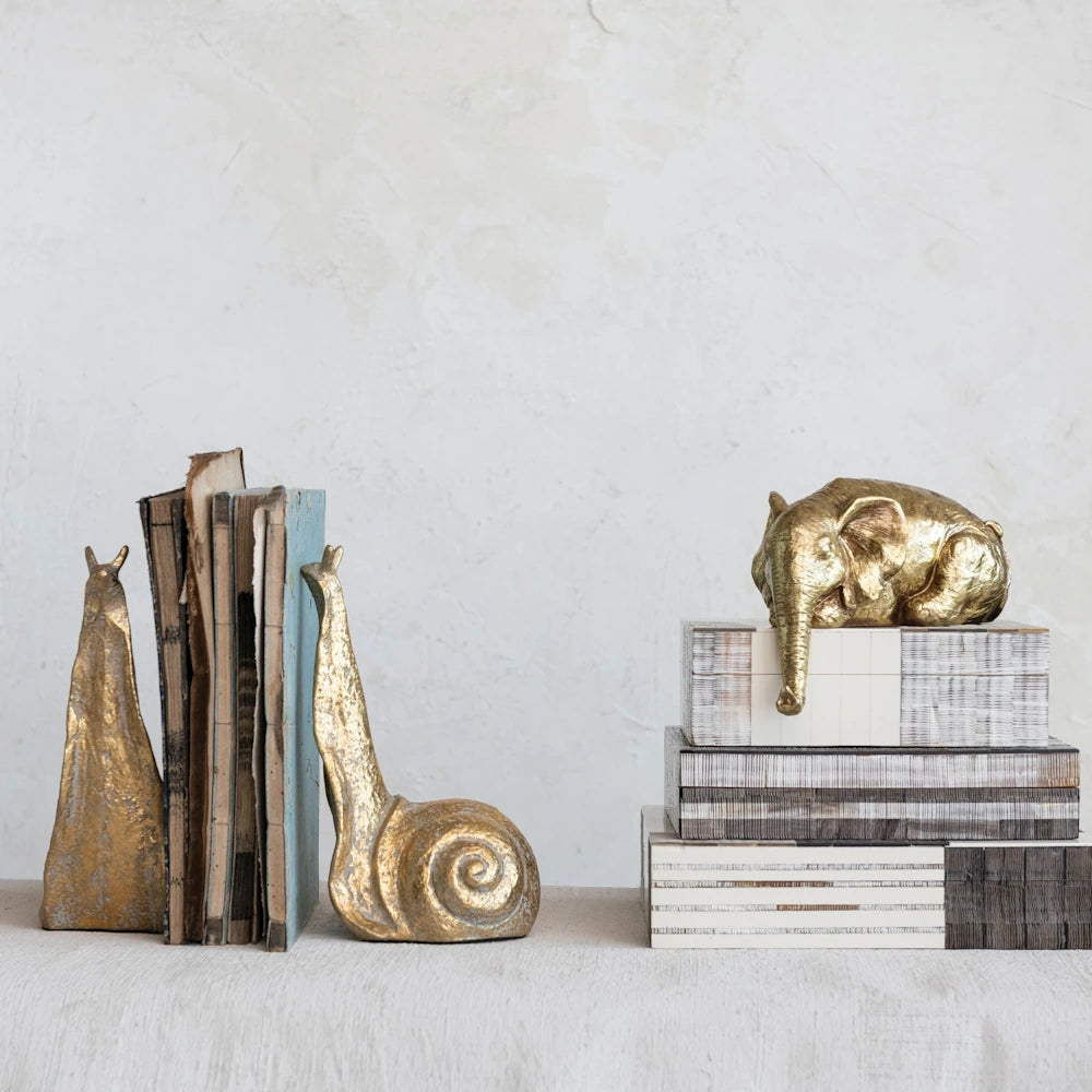 Brass snail bookends with old books and gold elephant home decor accents on textured white background.