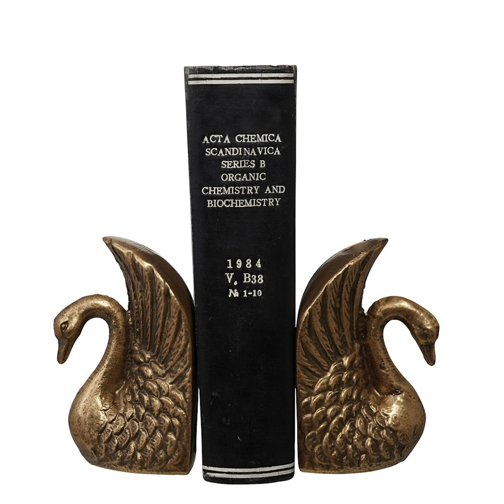 Pair of brass swan bookends with detailed feathers holding up a black vintage book against a white background.