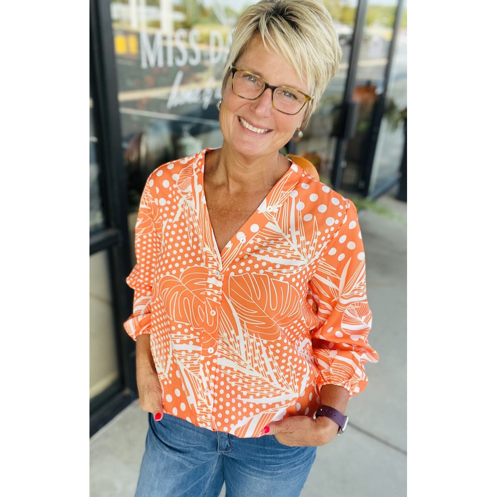 Smiling woman with short blonde hair wearing a bright orange blouse with white leaf and polka dot prints.