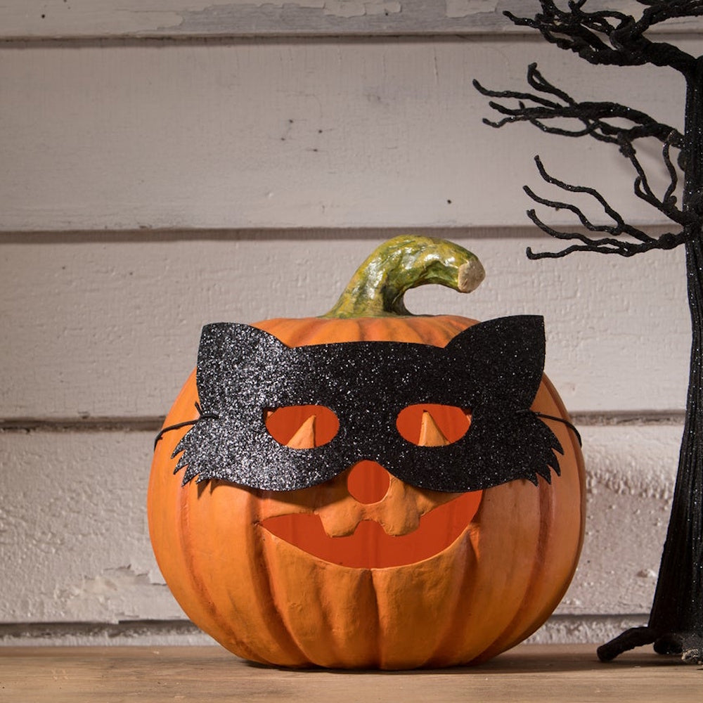 Carved orange pumpkin wearing a black glitter cat mask beside a spooky black tree.
