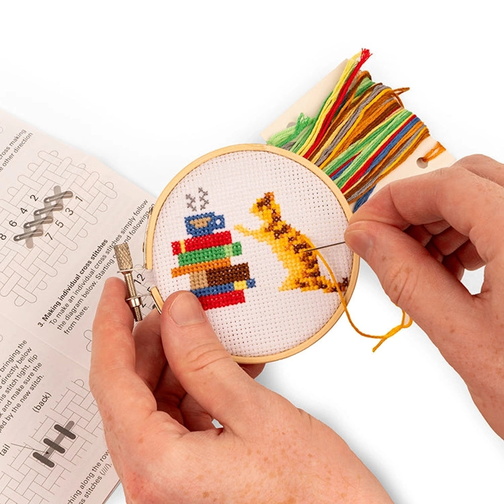 Hands stitching a mini cross-stitch kit featuring a ginger cat next to a stack of colorful books and a blue teacup.