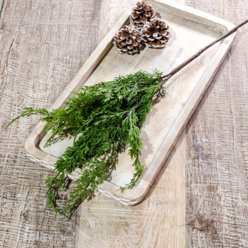 Artificial green cedar spray and pinecones arranged on a rectangular wooden tray.