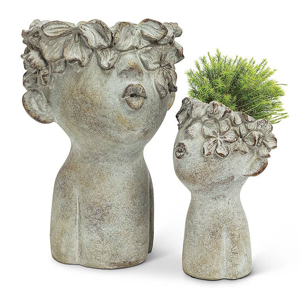 Two stone-look cement face planters with floral crowns and kissing lips, one holding a green plant.