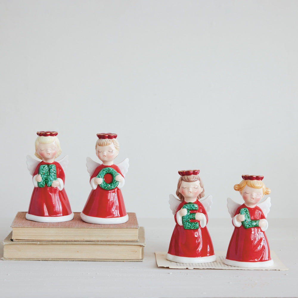 Four ceramic angel figurines in red dresses holding letter-shaped wreaths that spell NOEL on books.