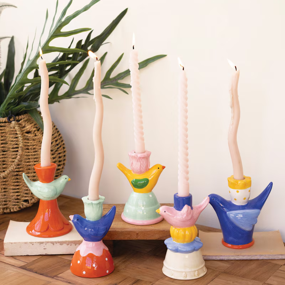 Five colorful, hand-painted ceramic bird taper candle holders with lit, wavy cream candles on a wooden surface.