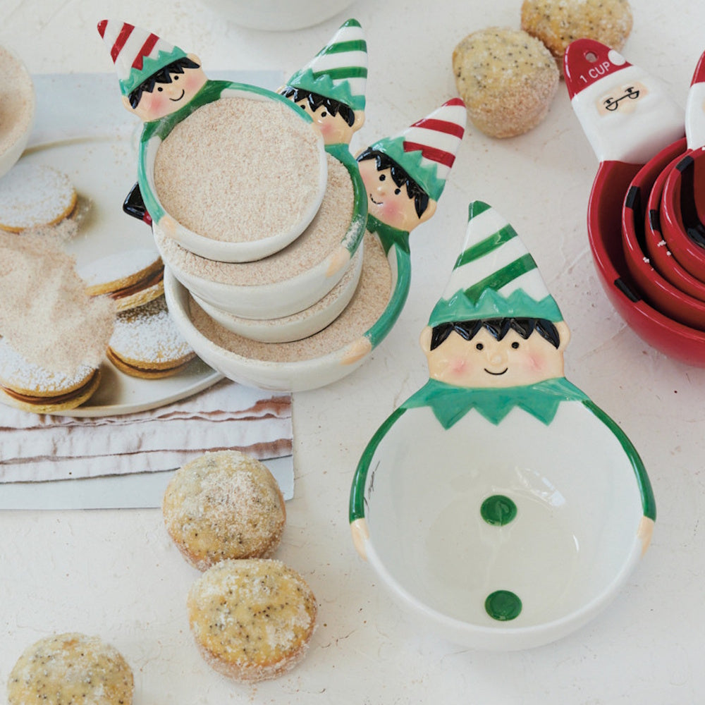 Elf-shaped measuring cups filled with dry ingredients, surrounded by baked treats on a kitchen counter.