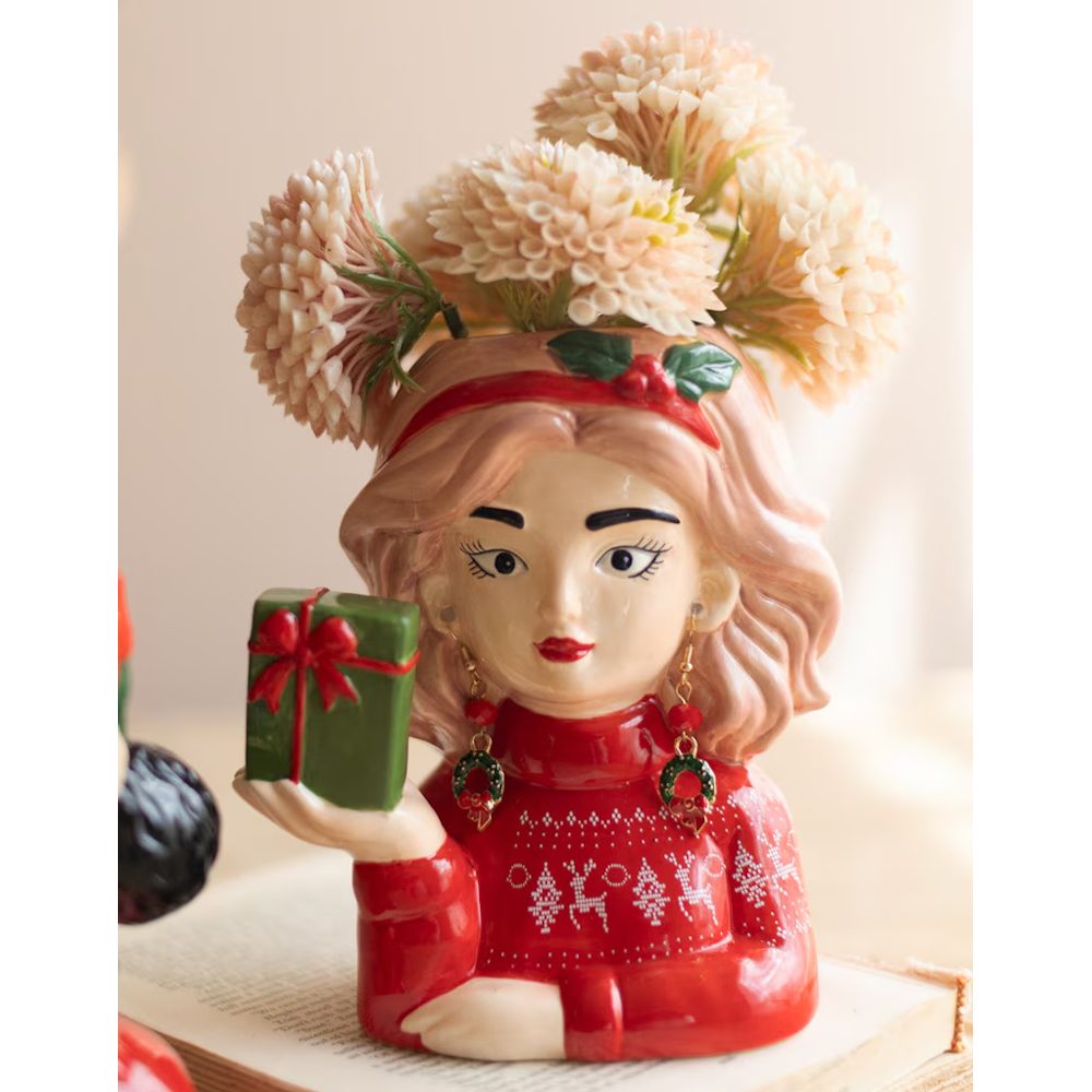 Ceramic woman head vase in a red holiday sweater holding a green gift box, filled with flowers.