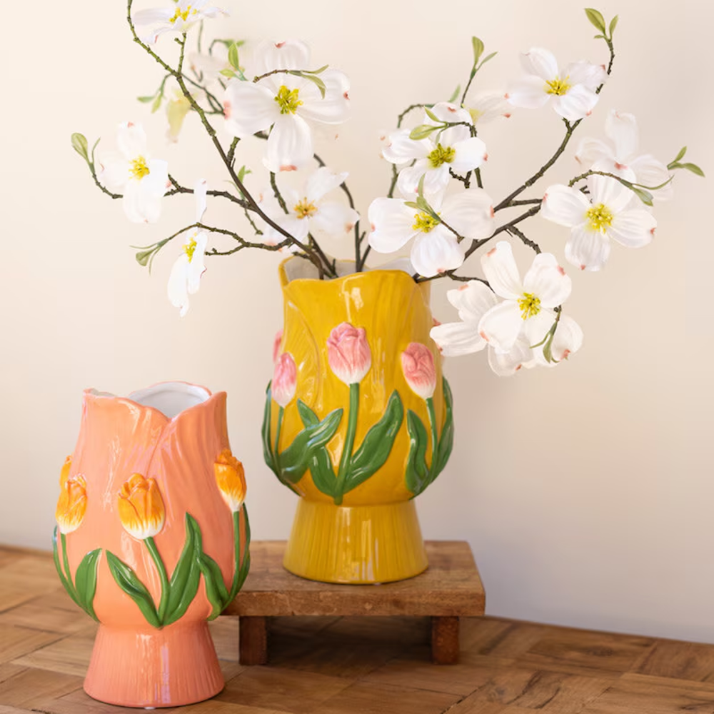 Two ceramic tulip-shaped vases in yellow and peach with 3D floral details, holding white dogwood branches on a wood table.
