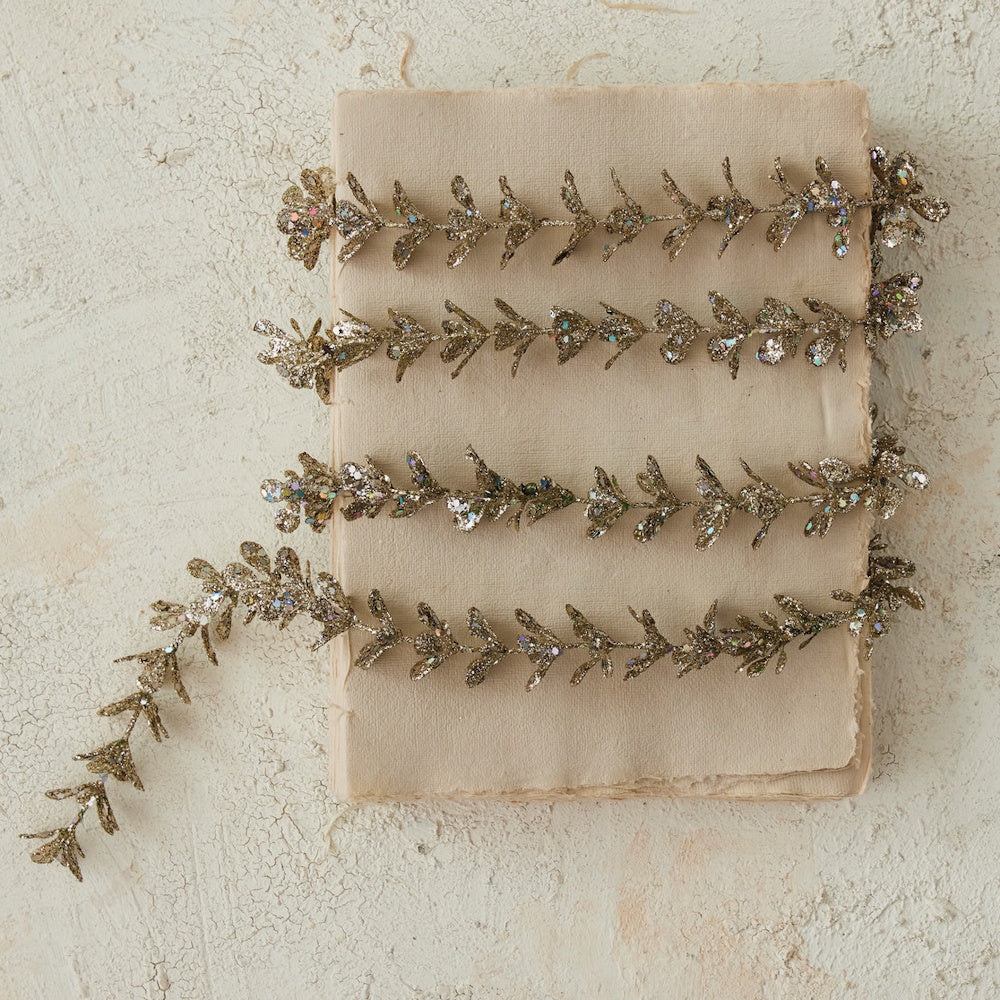 Champagne faux boxwood garland with glitter, coiled across a stack of textured neutral paper.