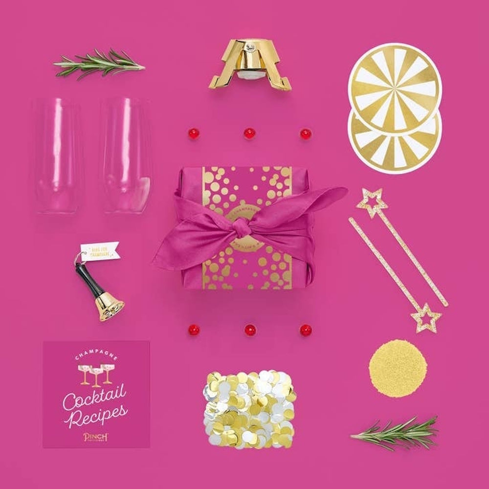 Champagne kit items including glasses, a recipe book, a stopper, and accessories on a bright pink background.