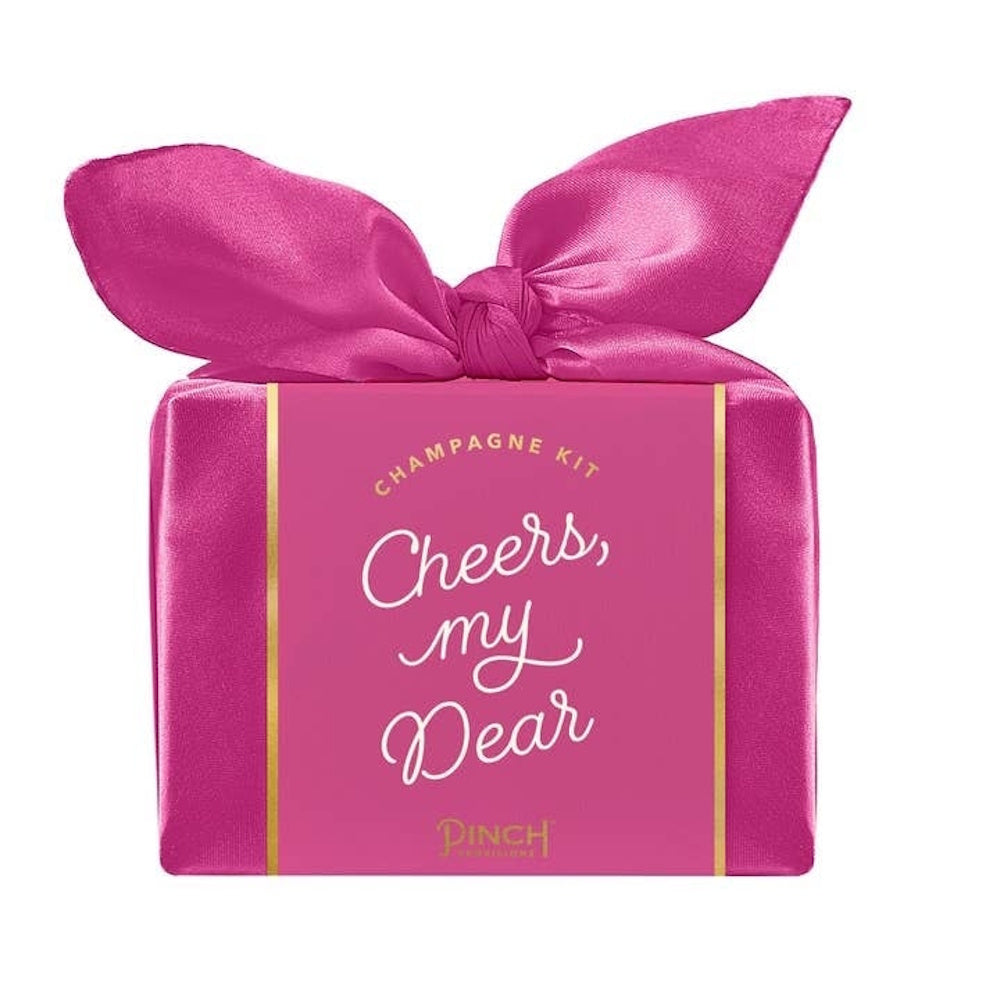 A bright pink champagne kit wrapped in satin fabric with a large bow, reading "Cheers, my Dear."