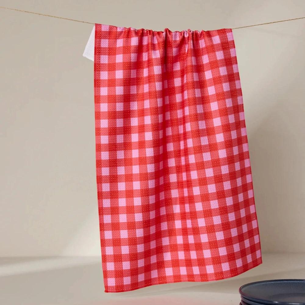 A red and pink gingham checkered kitchen towel hanging from a clothesline against a neutral background.