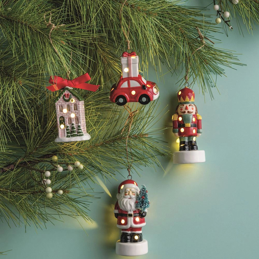Four LED lighted ceramic Christmas ornaments hanging on evergreen branches against a teal background.