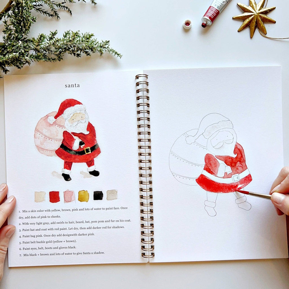 Open Christmas watercolor workbook showing a painted Santa Claus and a hand painting the outline.
