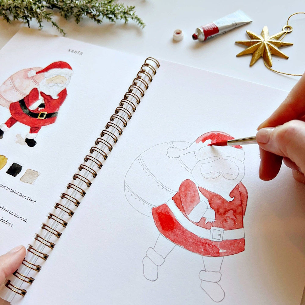 Close-up of a hand using a paintbrush to fill in a Santa Claus illustration in a watercolor book.