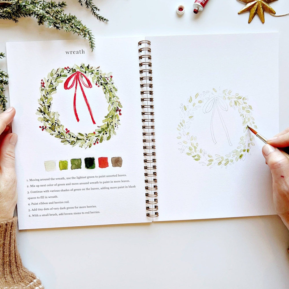 Open Christmas watercolor workbook with step-by-step instructions and a hand painting a wreath.