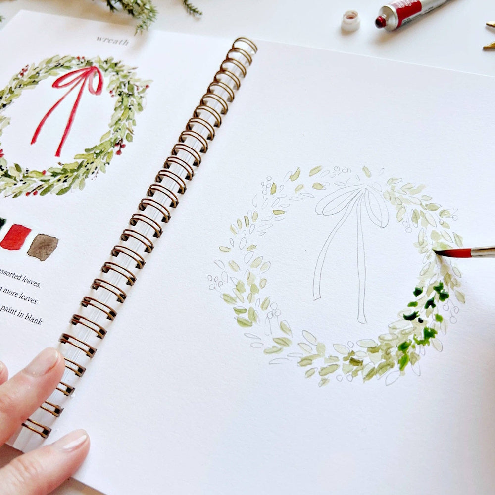 Close-up of a hand using a fine paintbrush to detail a holiday wreath outline in a watercolor book.