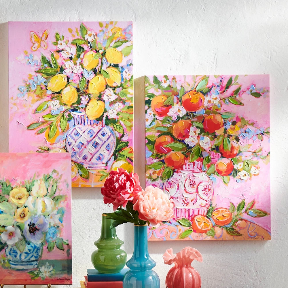 Three colorful, impressionist paintings of citrus bouquets in vases, displayed on a wall above real vases and flowers.