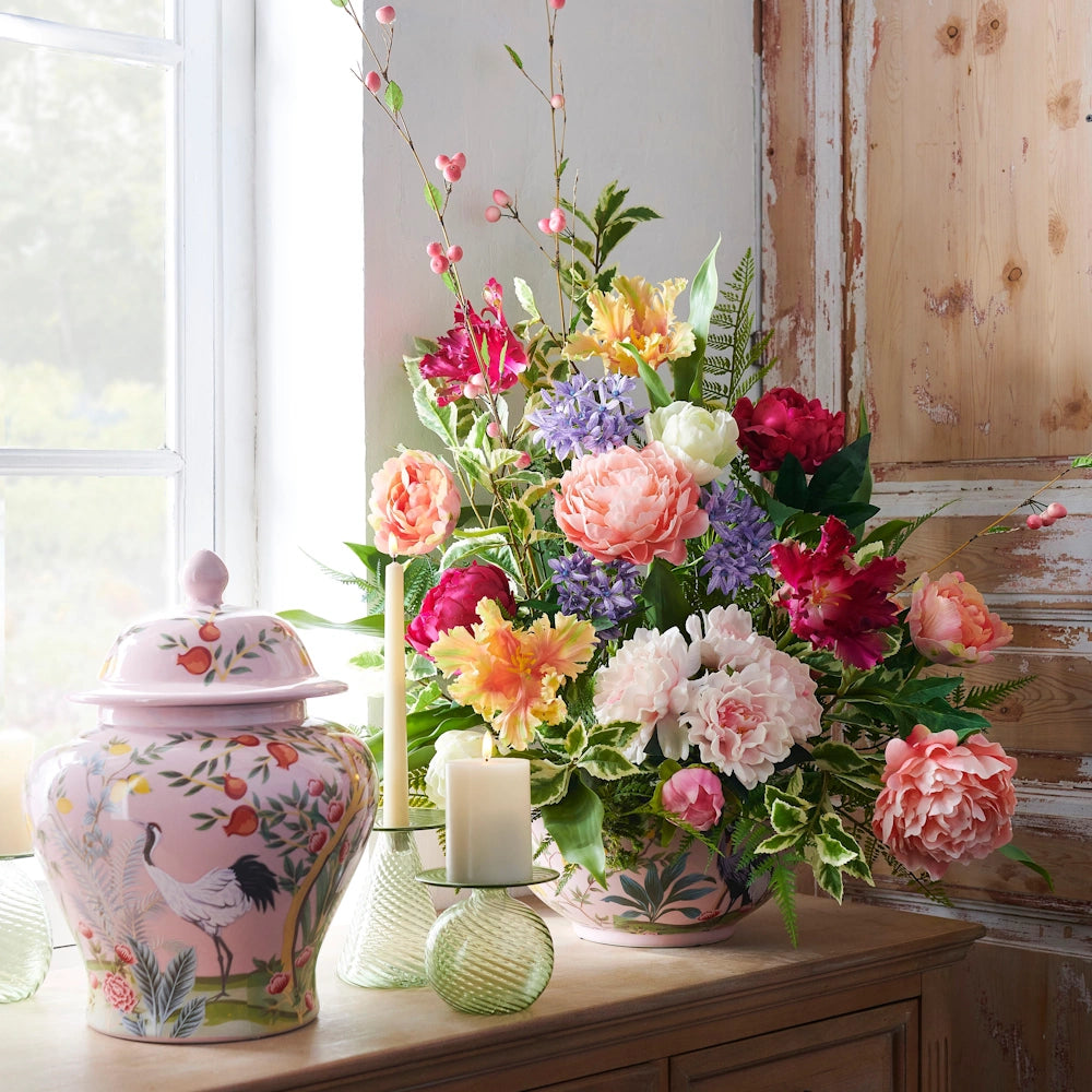 A pink floral arrangement and matching ginger jar with bird motifs sit on a wooden table next to lit candles.