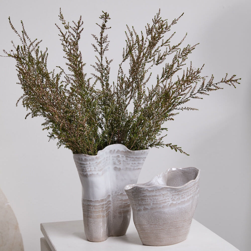 Two wavy, textured ceramic vases sit on a white surface, one holding a large bouquet of green sprigs.